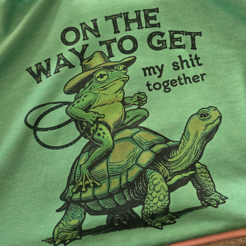 Light green t-shirt with a graphic of a frog riding a turtle and text, next to jeans and keys.