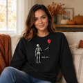 A woman sitting on a couch wearing a black crewneck sweatshirt with a graphic of a skeleton holding a red balloon and the text 'Well, shit.