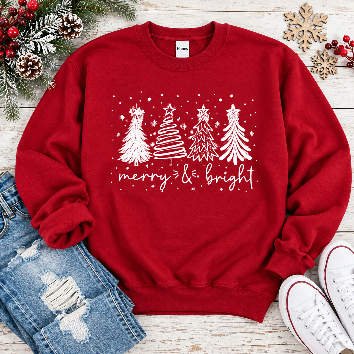 A red crewneck sweatshirt with a white Christmas tree graphic and 'merry & bright' text, styled in a flat lay with jeans, white sneakers, pinecones, and holiday decorations on a white wood background.