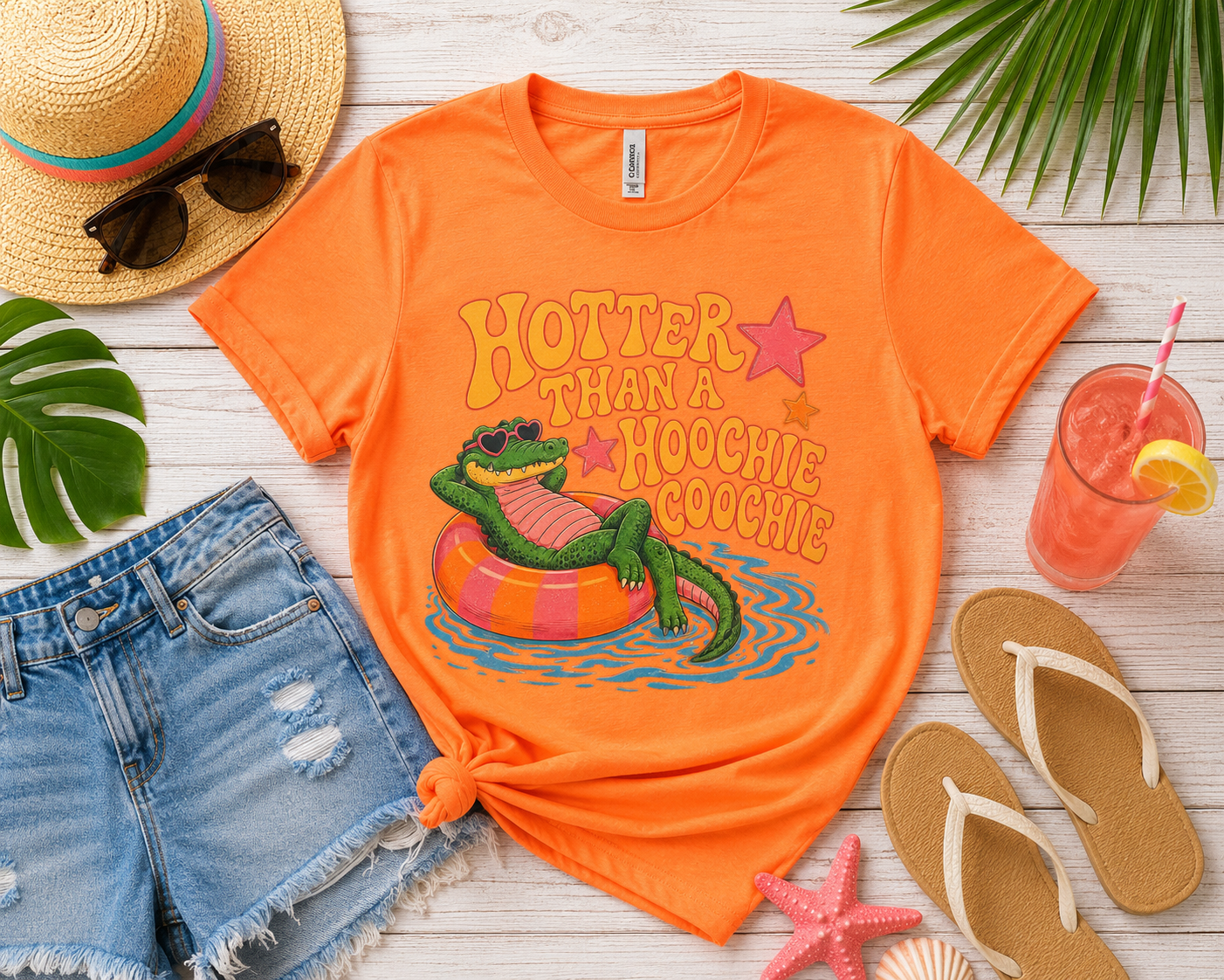 Orange t-shirt with a graphic of a crocodile in an inner tube and text, surrounded by beach accessories on a wooden surface.