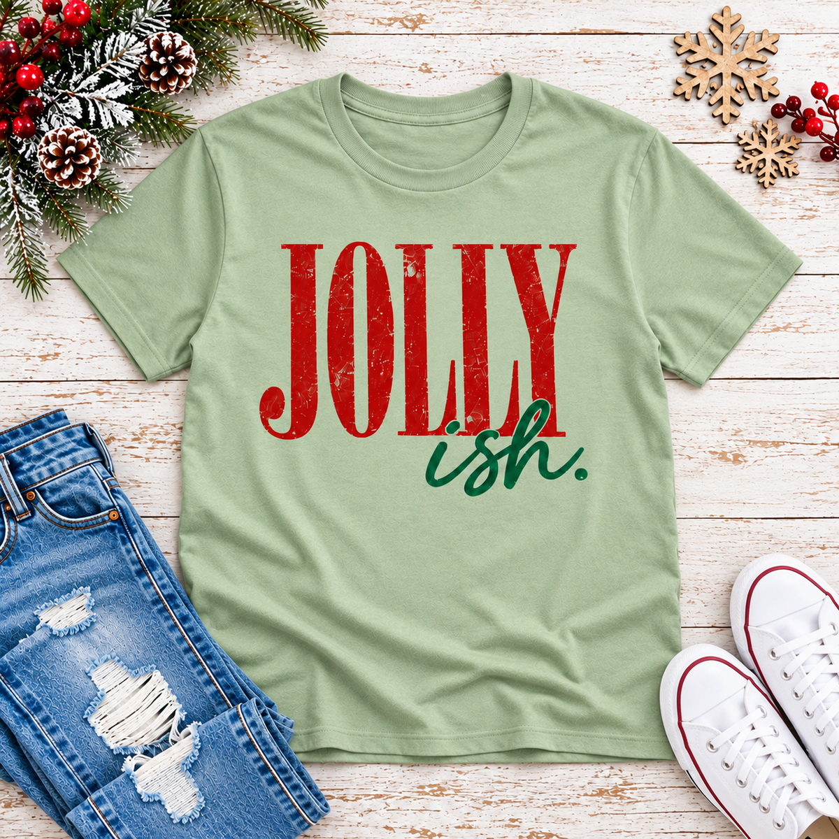 A light sage green t-shirt with a distressed red and green graphic reading 'JOLLY ish.' laid flat on a white wood background, styled with jeans, white sneakers, pinecones, and holiday greenery.