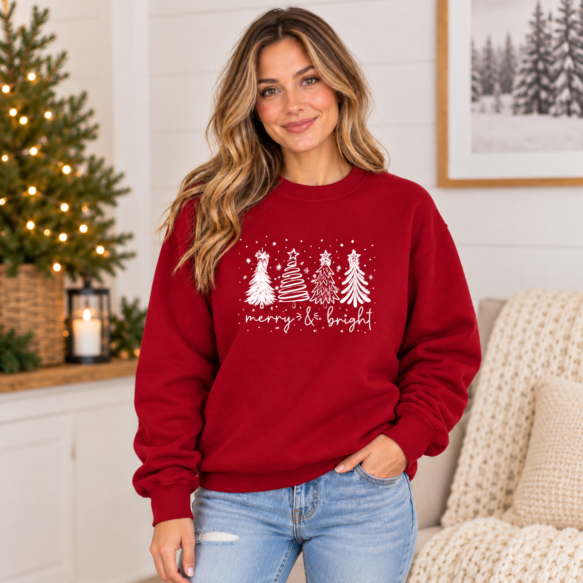 A woman wearing a festive red crewneck sweatshirt with a white graphic of Christmas trees and the text 'merry & bright' in a cozy living room setting.