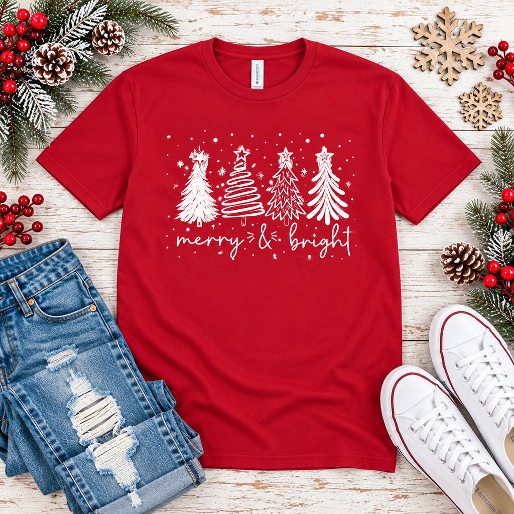 A red short-sleeve crewneck t-shirt with a white graphic of three Christmas trees and the text 'merry & bright', styled in a flat lay with jeans, white sneakers, pinecones, and holiday greenery on a whitewashed wood background.