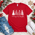 A red short-sleeve crewneck t-shirt with a white graphic of three Christmas trees and the text 'merry & bright', styled in a flat lay with jeans, white sneakers, pinecones, and holiday greenery on a whitewashed wood background.