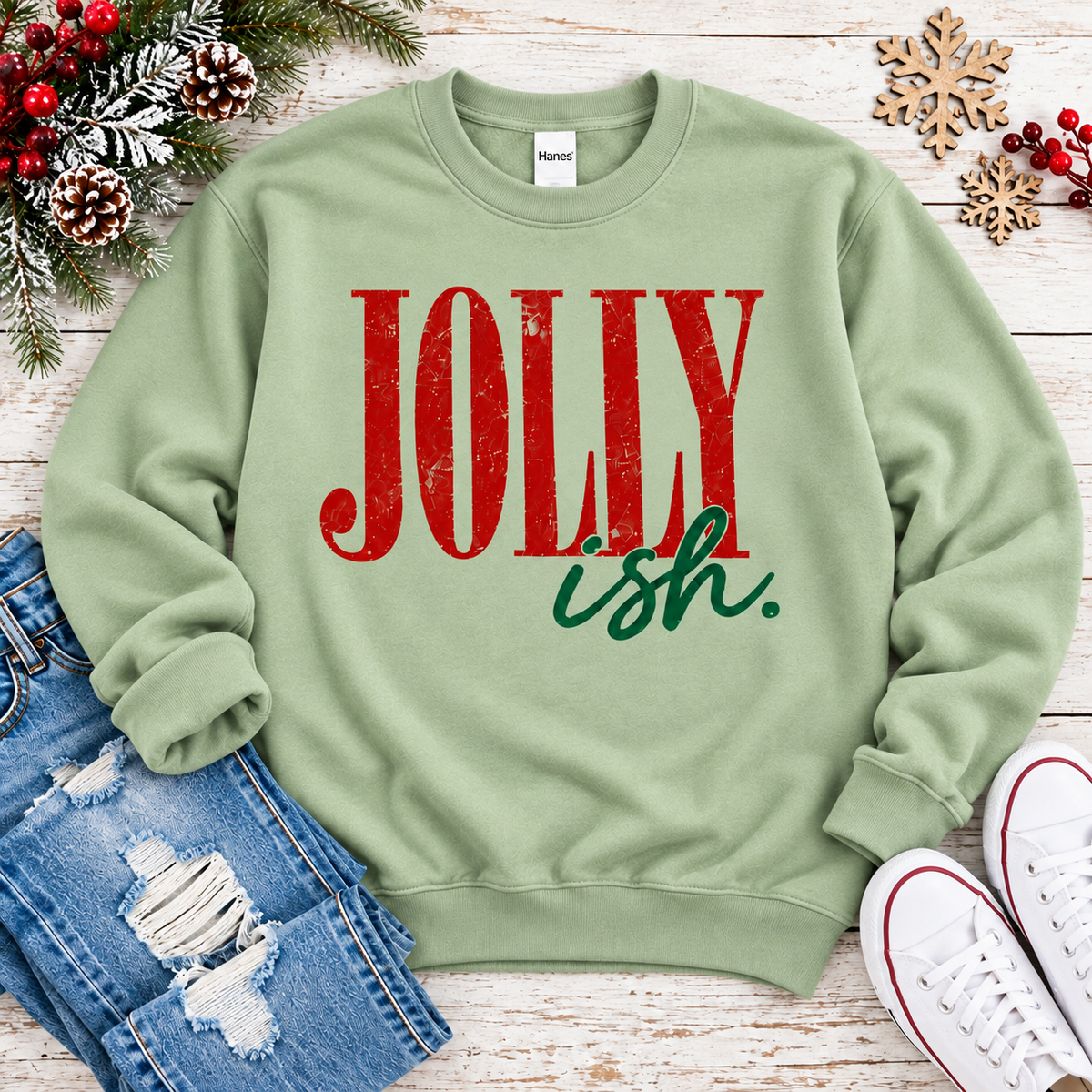 A sage green crewneck sweatshirt with 'JOLLY ish.' printed on the front in distressed red and dark green lettering, styled in a flat lay with blue jeans, white sneakers, and holiday decorations on a white wood background.