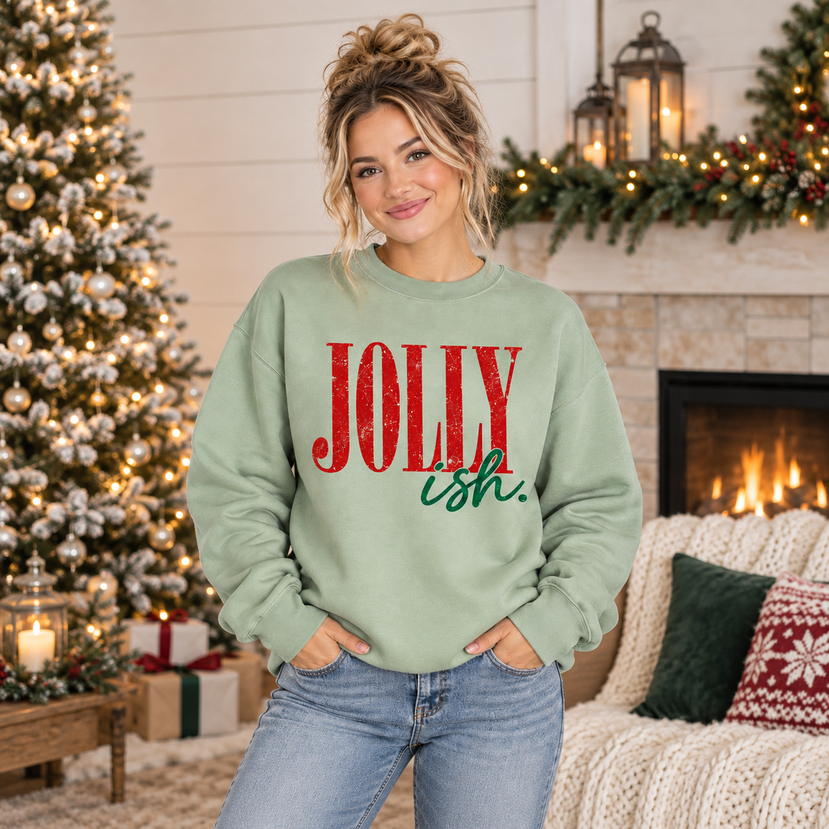 A woman wearing a sage green crewneck sweatshirt with 'JOLLY ish.' printed on the front, posed in a cozy holiday living room setting with a decorated Christmas tree and fireplace in the background.