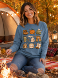 A woman sitting by a campfire outdoors wearing a blue crewneck sweatshirt with a cute cartoon graphic of autumn and winter elements, paired with ripped blue jeans.