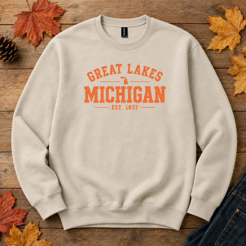 A cream-colored crewneck sweatshirt with an orange graphic reading 'GREAT LAKES MICHIGAN EST. 1837' laid flat on a rustic wooden surface with autumn leaves and jeans as props.