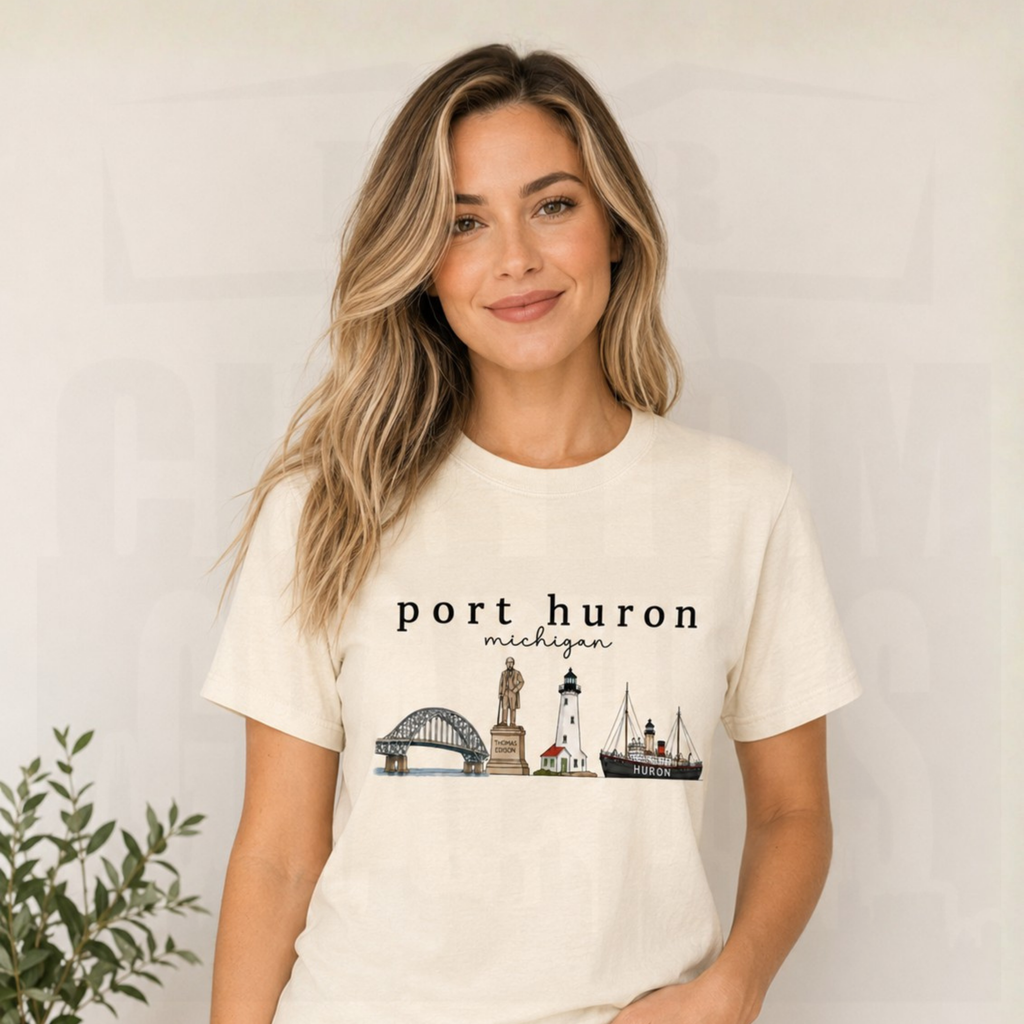 A woman with blonde-highlighted hair wearing a cream-colored t-shirt with a 'port huron michigan' graphic featuring lighthouses and a ship, posed against a light gray wall with a small plant in the corner.
