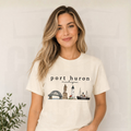 A woman with blonde-highlighted hair wearing a cream-colored t-shirt with a 'port huron michigan' graphic featuring lighthouses and a ship, posed against a light gray wall with a small plant in the corner.