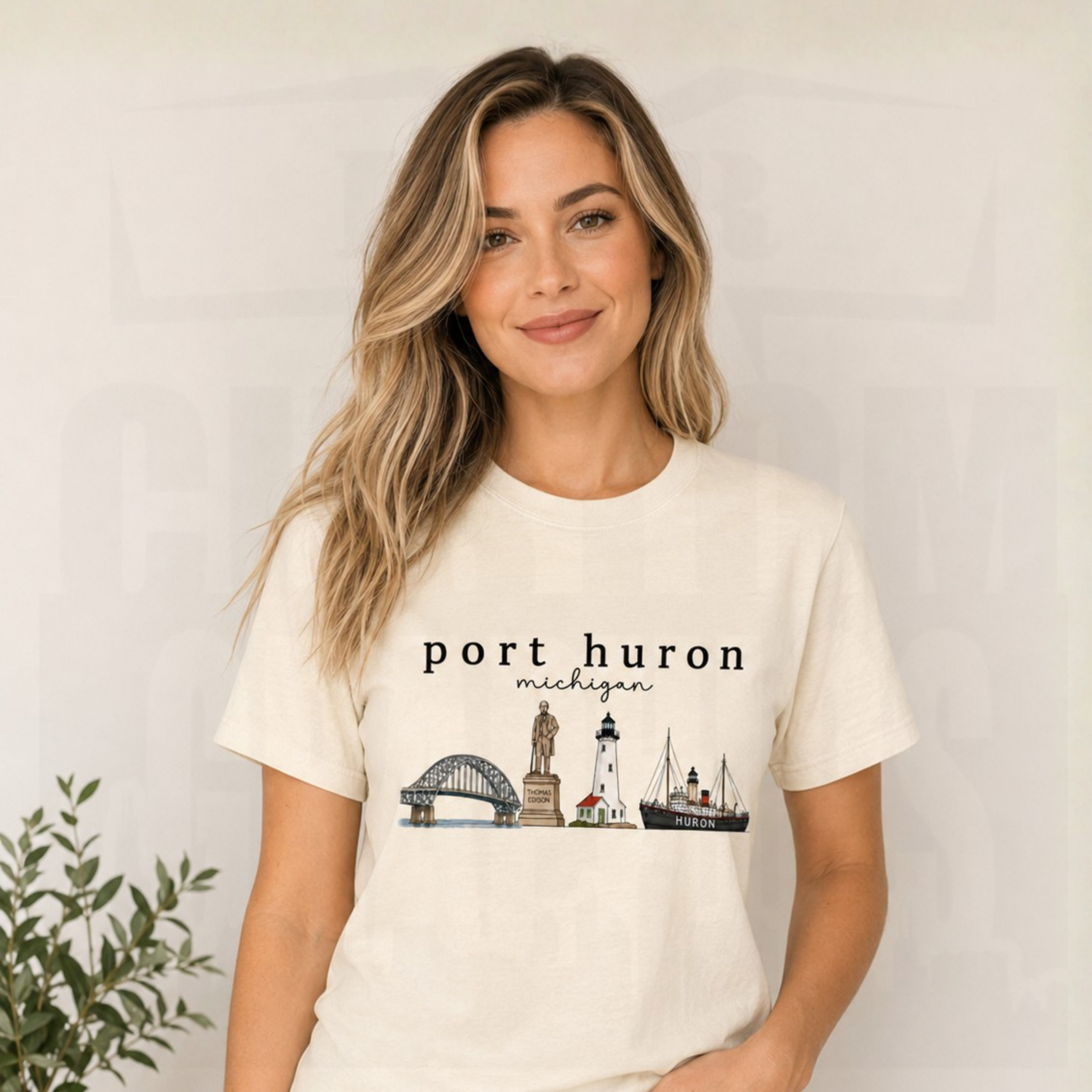 A woman with blonde-highlighted hair wearing a cream-colored t-shirt with a 'port huron michigan' graphic featuring lighthouses and a ship, posed against a light gray wall with a small plant in the corner.