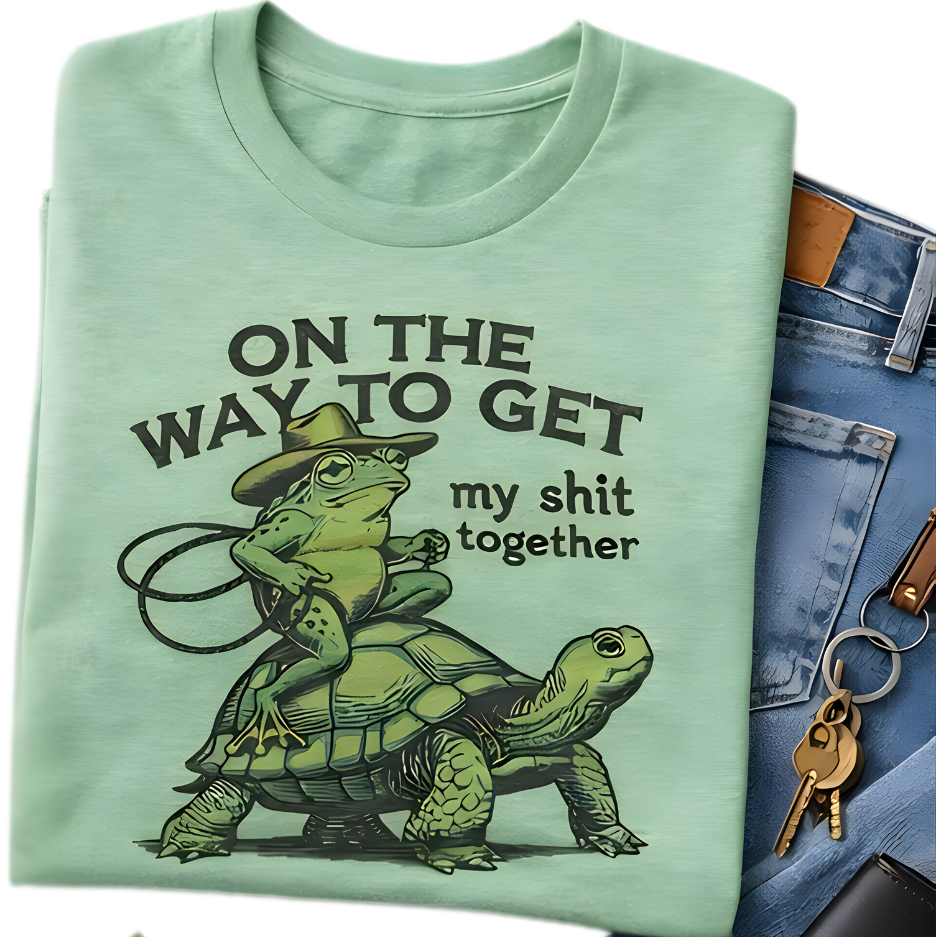 Light green t-shirt with a graphic of a frog riding a turtle and text, next to jeans and keys.