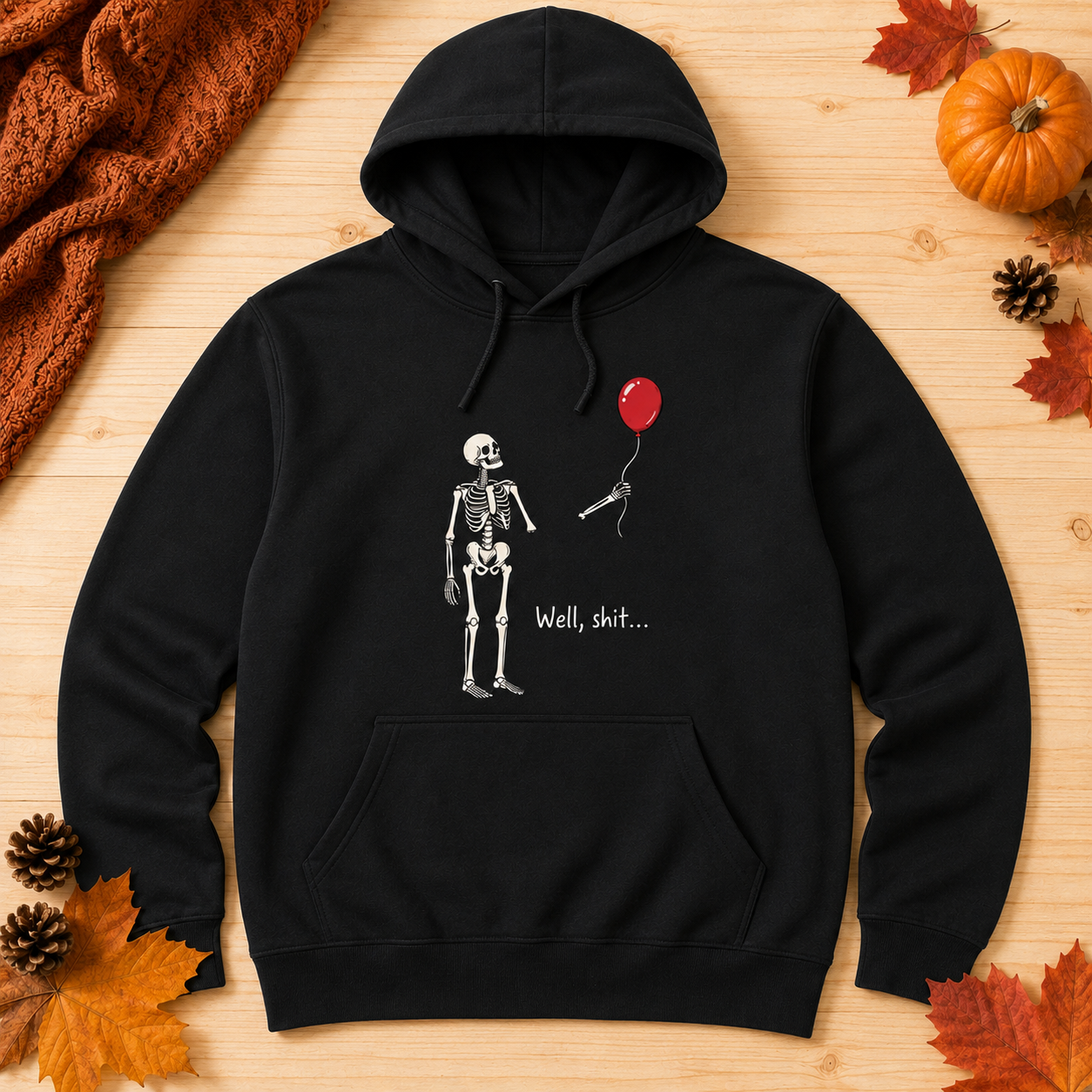 A black pullover hoodie laid flat on a light wood surface, featuring a white skeleton graphic holding a red balloon with the text 'Well, shit.