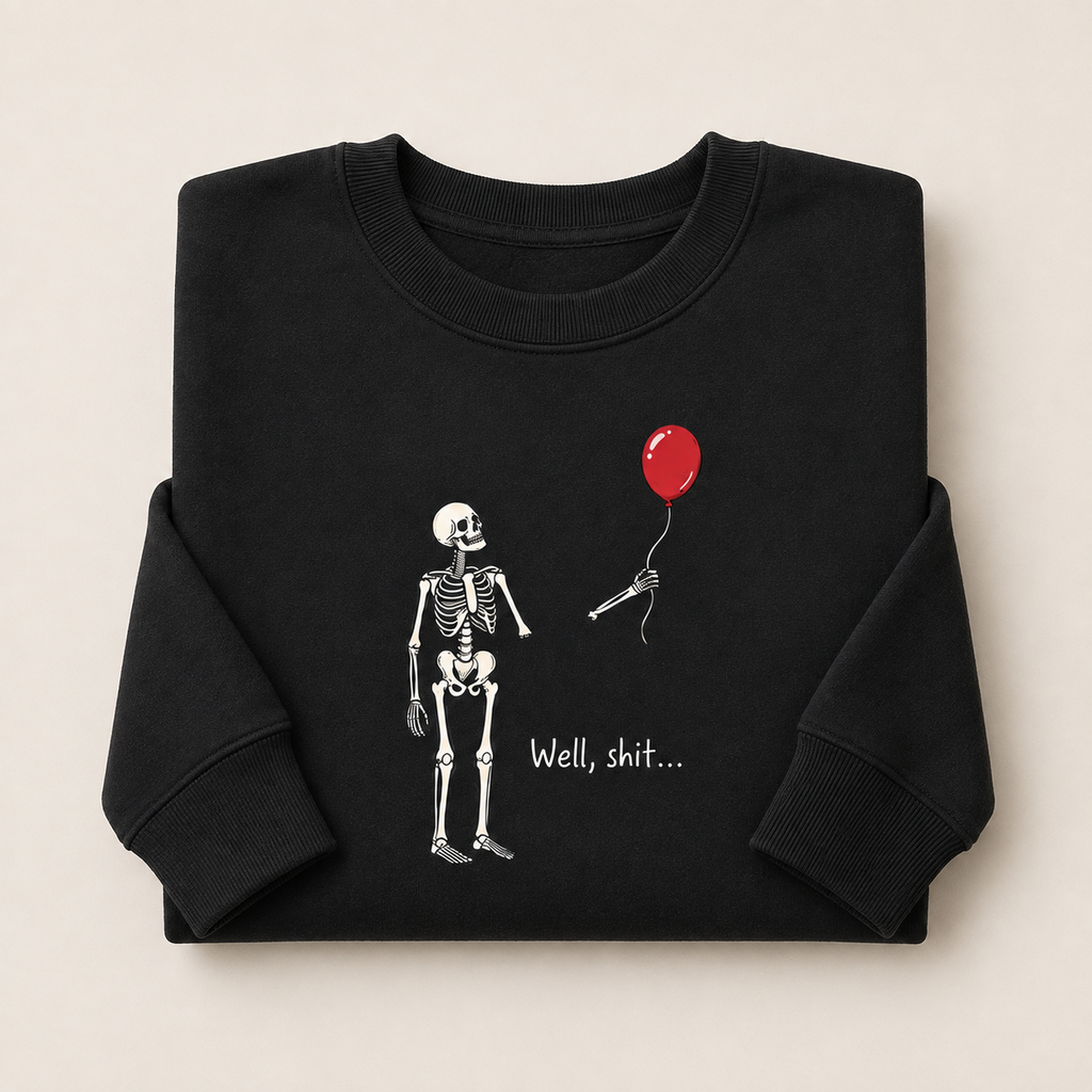 A folded black crewneck sweatshirt on a light beige background, featuring a graphic of a white skeleton holding a red balloon with the text 'Well, shit.